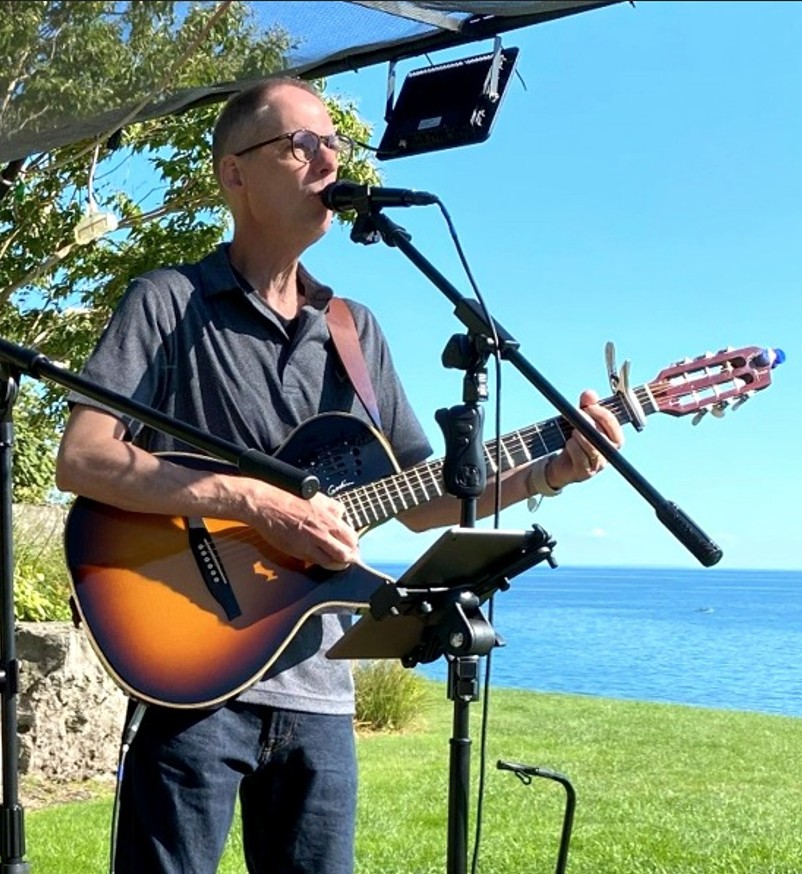 Jon singing into a microphone with guitar, water visible behind him